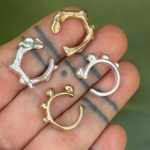 May include: Four ear cuffs in gold and silver tones. Two cuffs feature a smooth, curved band with small spherical accents along the outer edge. The other two cuffs have an irregular, textured, organic shape resembling a twig or branch.