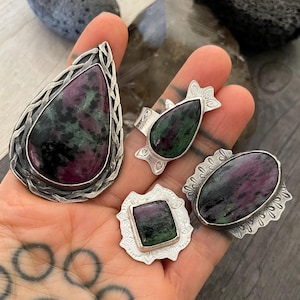 May include: Four silver rings with ruby in zoisite stones are displayed. The rings have different shapes: a teardrop, a rounded oval, a square, and a smaller teardrop. The stones feature a mix of deep red, green, and black colors.