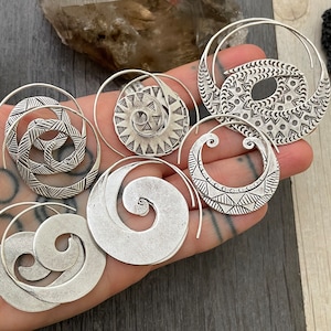 May include: A collection of silver-toned spiral earrings featuring various etched geometric patterns, including circular, crescent, and swirling designs. These metallic earrings have a polished finish and are displayed together in a group.