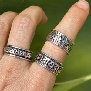 May include: Three silver-toned metal rings worn on fingers. Two rings feature embossed script lettering around the band, while the third ring displays an intricate, openwork filigree pattern with a braided border design along the top and bottom edges.