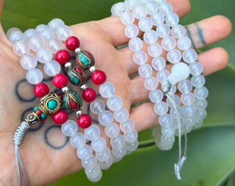 May include: Two multi-strand bracelets made of translucent white beads. One bracelet features accents of bright red spherical beads and ornate metal spacers inlaid with turquoise and red stones. Both are finished with grey cord knots.