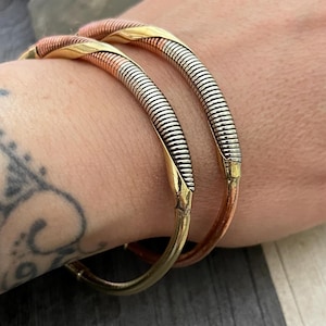 Op de afbeelding: Twee zilveren en koperen armbandjes met een gedraaid ontwerp. De armbandjes worden om de pols gedragen.