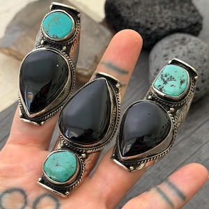 May include: Three silver rings with large, teardrop-shaped black stones and turquoise accents. The rings are ornate, with detailed metalwork around the stones. The rings are displayed on a hand.