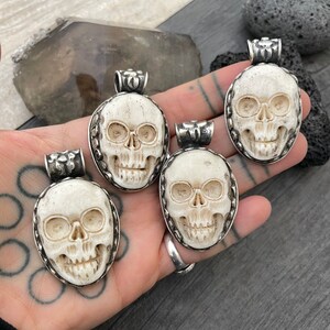 May include: Four white bone skull pendants with silver accents. Each pendant features a detailed carving of a skull with eye sockets, a nose, and a mouth.