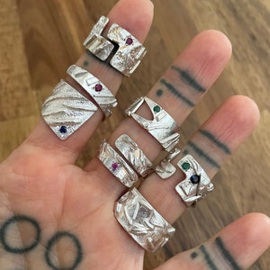 May include: A set of six silver rings with different designs. Each ring features a unique textured surface and a small gemstone. The gemstones are red, green, and blue.