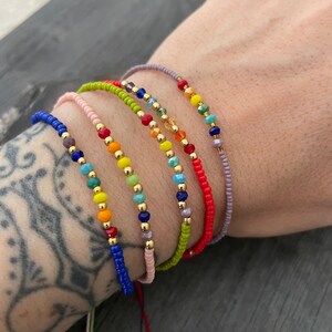 May include: A set of colorful beaded bracelets with gold accents. The bracelets are layered on a person's wrist and feature a variety of colors, including red, orange, yellow, green, blue, and purple.