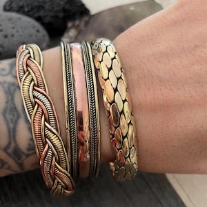 Puede incluir: Tres pulseras apiladas hechas de metal tejido. La pulsera superior tiene una textura escamosa, la pulsera del medio es una banda de cobre con un borde decorativo, y la pulsera inferior es un diseño trenzado con tonos cobre, plata y oro.