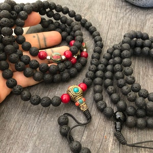 May include: Two long necklaces made of black porous lava stone beads. One necklace features a central brass-toned metal bead with turquoise and red accents, flanked by red beads. The other necklace has a single smooth black bead at the end.