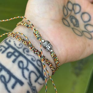 May include: A multi-colored braided cord bracelet featuring a silver-toned metal Buddha head charm. The bracelet is constructed from thin, interwoven threads in shades of yellow, red, green, and black, and includes adjustable sliding knots.
