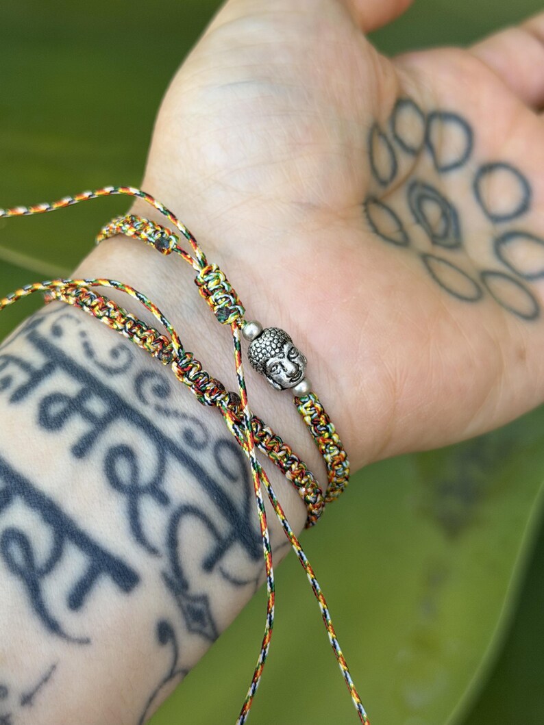May include: A multi-colored braided cord bracelet featuring a silver-toned metal Buddha head charm. The bracelet is constructed from thin, interwoven threads in shades of yellow, red, green, and black, and includes adjustable sliding knots.