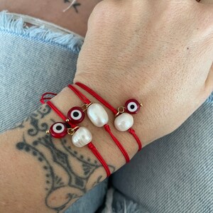 K&ouml;nnte beinhalten: Drei rote Armb&auml;nder mit wei&szlig;en Perlenanh&auml;ngern und b&ouml;sen Augenanh&auml;ngern. Die Armb&auml;nder sind um ein Handgelenk gebunden.