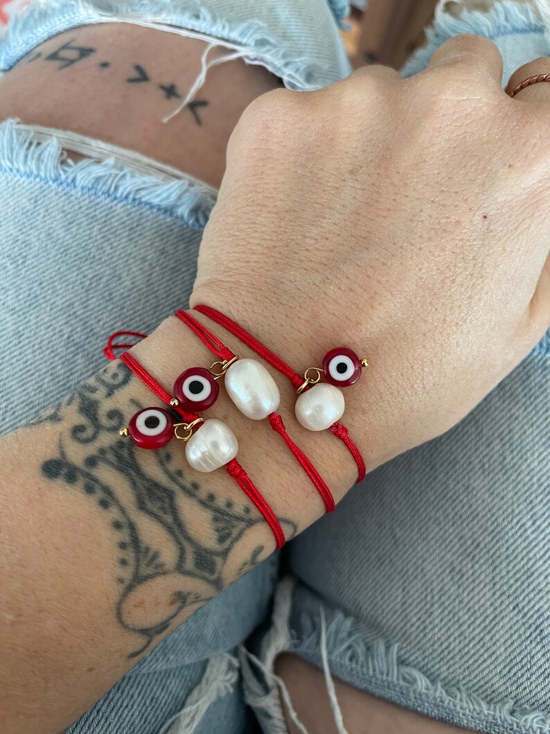 K&ouml;nnte beinhalten: Drei rote Armb&auml;nder mit wei&szlig;en Perlenanh&auml;ngern und b&ouml;sen Augenanh&auml;ngern. Die Armb&auml;nder sind um ein Handgelenk gebunden.