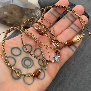 May include: Two adjustable bracelets made of braided multi-colored cord in shades of green, yellow, and red. One bracelet features a gold-toned Buddha charm and a brown rudraksha bead, while the other displays a silver-toned eternal knot charm.