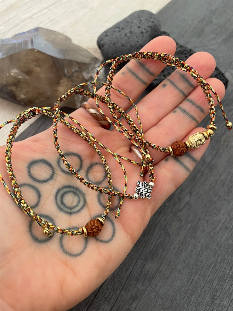 May include: Two adjustable bracelets made of braided multi-colored cord in shades of green, yellow, and red. One bracelet features a gold-toned Buddha charm and a brown rudraksha bead, while the other displays a silver-toned eternal knot charm.