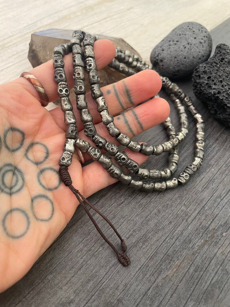 May include: A silver skull bead necklace with a brown cord tie. The necklace is made of multiple strands of beads and is approximately 30 inches long.