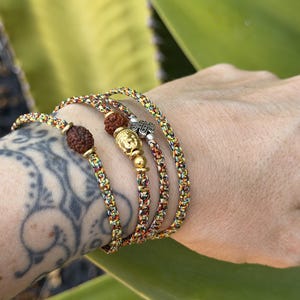 May include: A multi-strand bracelet made of braided, multicolored cord featuring two dark brown, textured rudraksha beads, a gold-toned Buddha head charm, and a small silver-toned metal charm with an intricate, embossed design.