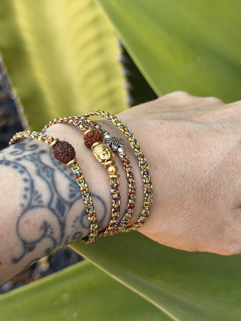 May include: A multi-strand bracelet made of braided, multicolored cord featuring two dark brown, textured rudraksha beads, a gold-toned Buddha head charm, and a small silver-toned metal charm with an intricate, embossed design.