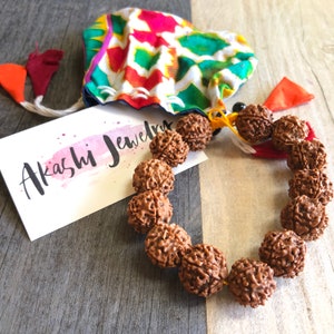May include: A brown wooden mala bracelet with 18 beads. The bracelet is tied with a yellow and orange string. The bracelet is on a wooden surface with a colorful fabric pouch and a card that says "Akashi Jewelry".