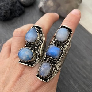 May include: Two silver rings with three iridescent moonstone gemstones each. The rings are designed with intricate details and have a curved shape.