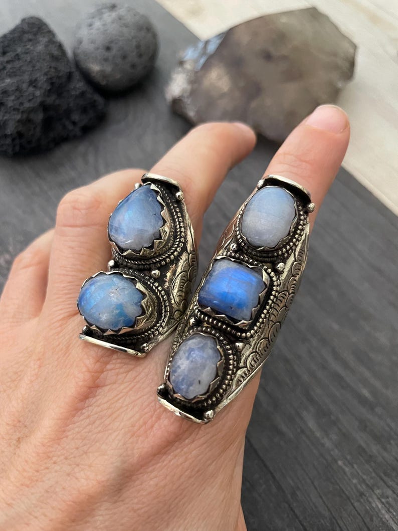 May include: Two silver rings with three iridescent moonstone gemstones each. The rings are designed with intricate details and have a curved shape.