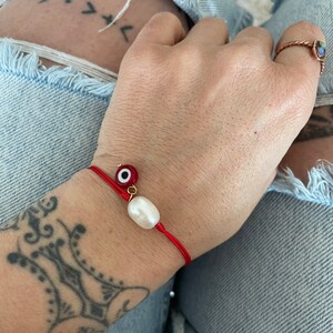 K&ouml;nnte beinhalten: Rotes Armband mit einem wei&szlig;en Perlenanh&auml;nger und einem blauen und goldenen Gl&uuml;cksbringer.