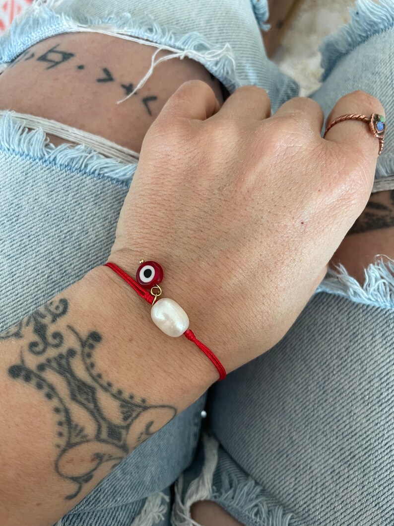 K&ouml;nnte beinhalten: Rotes Armband mit einem wei&szlig;en Perlenanh&auml;nger und einem blauen und goldenen Gl&uuml;cksbringer.