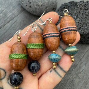 May include: Handmade wooden earrings with silver hooks. Each earring features a wooden barrel shape with colorful accents, including green, coral, and blue details. The earrings are finished with a dark blue or turquoise bead.
