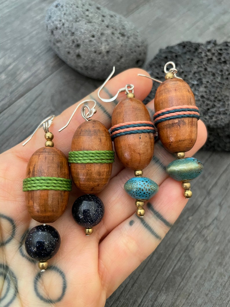 May include: Handmade wooden earrings with silver hooks. Each earring features a wooden barrel shape with colorful accents, including green, coral, and blue details. The earrings are finished with a dark blue or turquoise bead.