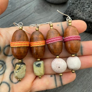 May include: Four handmade earrings with wooden beads, wrapped with orange and pink thread. The earrings have different stone and bead accents, including a light green stone and white stone beads. The earrings are on silver hooks.