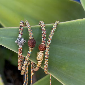 May include: A set of four braided bracelets made from multicolored thread. One bracelet features a silver-toned eternal knot charm, another includes a brown textured bead, and a third displays a gold-toned Buddha head bead with a brown bead.