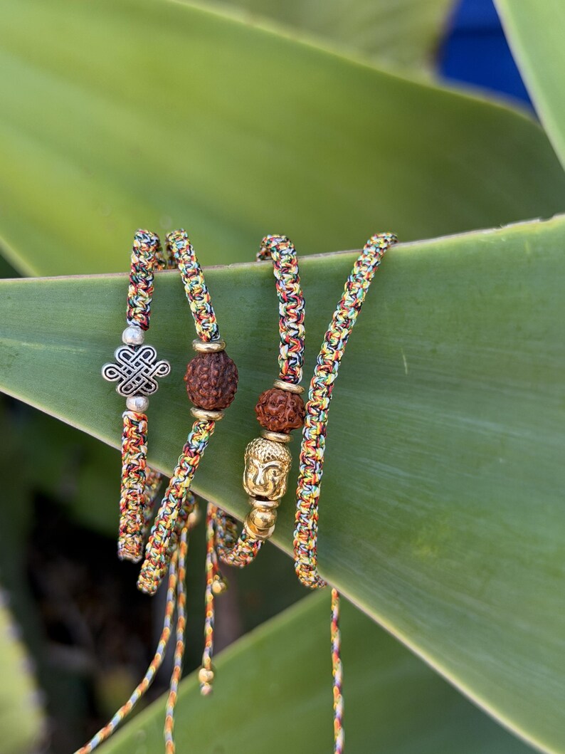 May include: A set of four braided bracelets made from multicolored thread. One bracelet features a silver-toned eternal knot charm, another includes a brown textured bead, and a third displays a gold-toned Buddha head bead with a brown bead.