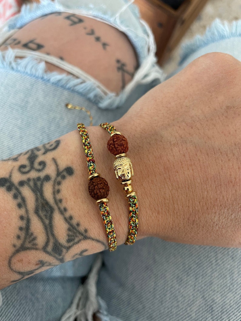 May include: Two bracelets featuring braided cords in yellow, green, and red. Each bracelet includes a round, textured brown rudraksha bead and gold-toned metal accents. One bracelet features a small gold-toned Buddha head charm as a centerpiece.