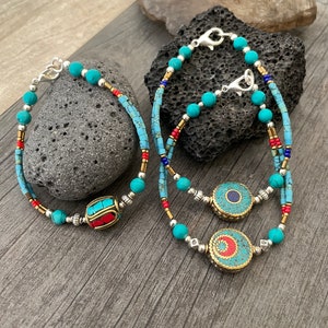Puede incluir: Tres pulseras de cuentas turquesa con cierres plateados. Una pulsera tiene un dije en forma de barril rojo y azul, y las otras dos tienen dijes redondos turquesa y dorados.