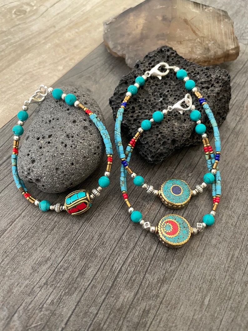 Puede incluir: Tres pulseras de cuentas turquesa con cierres plateados. Una pulsera tiene un dije en forma de barril rojo y azul, y las otras dos tienen dijes redondos turquesa y dorados.