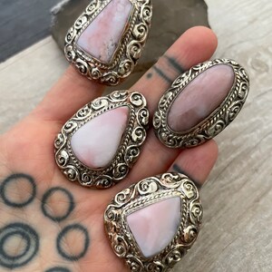 May include: Three silver rings with pink opal stones set in ornate filigree designs. The rings are on a hand with a tattoo.