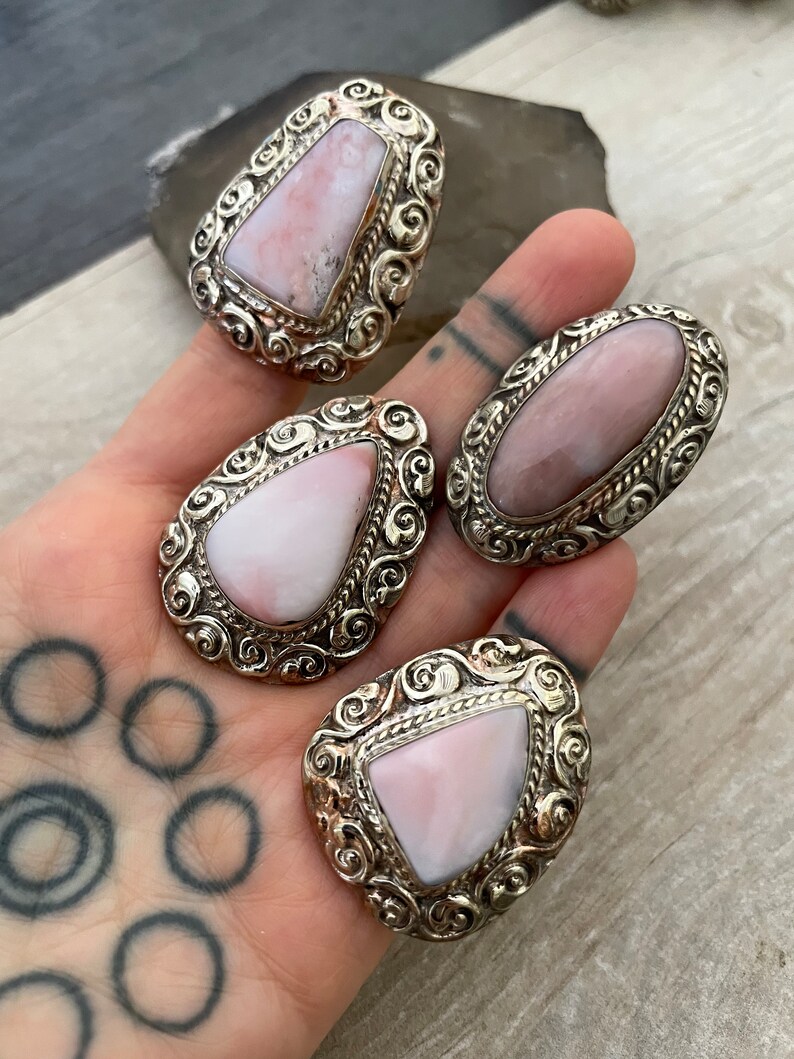 May include: Three silver rings with pink opal stones set in ornate filigree designs. The rings are on a hand with a tattoo.