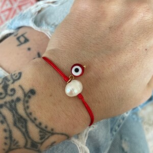 K&ouml;nnte beinhalten: Rotes Armband mit einem wei&szlig;en Perlenanh&auml;nger und einem blauen und roten b&ouml;sen Augenanh&auml;nger.