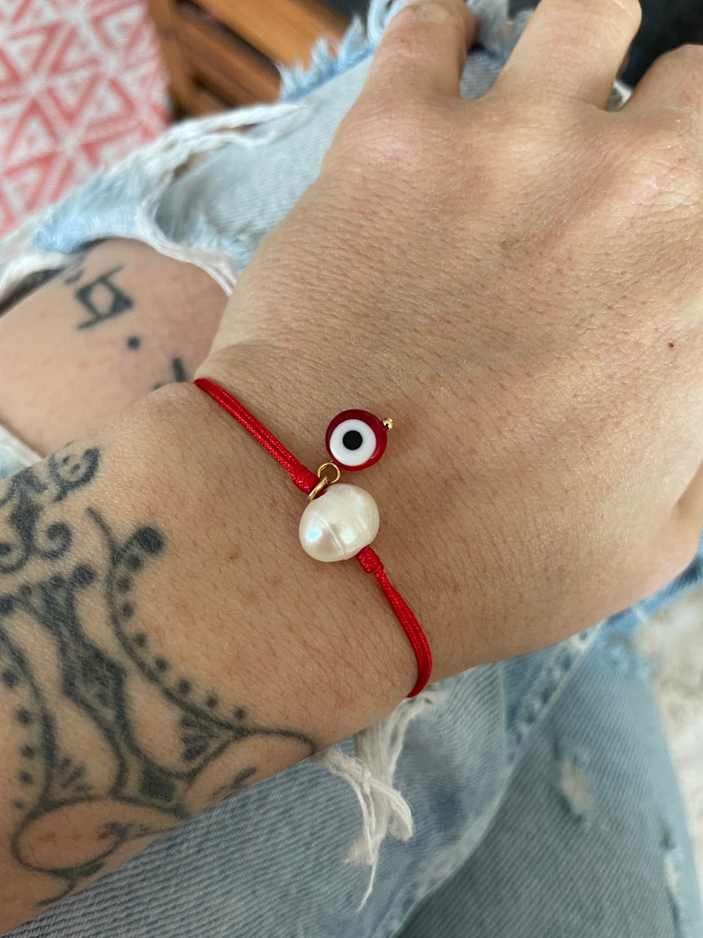 K&ouml;nnte beinhalten: Rotes Armband mit einem wei&szlig;en Perlenanh&auml;nger und einem blauen und roten b&ouml;sen Augenanh&auml;nger.