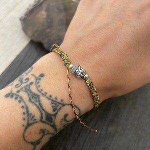 May include: A braided bracelet made of multicolored cord in shades of yellow, orange, and green. The bracelet features a central silver-toned metal charm shaped like a Buddha head, flanked by two small silver-toned metal beads on either side.