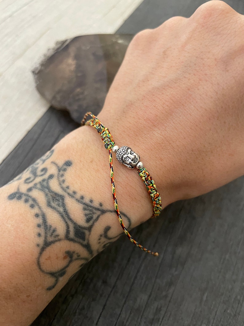May include: A braided bracelet made of multicolored cord in shades of yellow, orange, and green. The bracelet features a central silver-toned metal charm shaped like a Buddha head, flanked by two small silver-toned metal beads on either side.