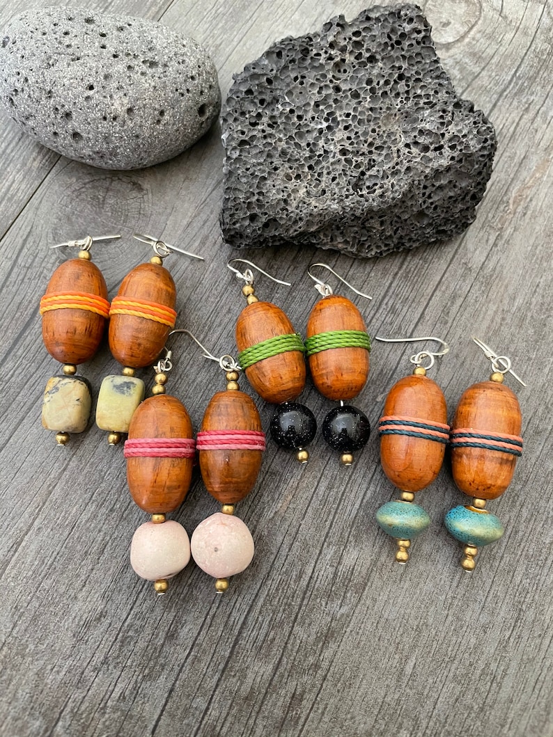 May include: A collection of handcrafted earrings with wooden and stone beads. The earrings feature various designs, including wooden oval beads wrapped with orange, green, pink, and black twine. Some earrings have stone or ceramic beads in white, black, and blue-green.