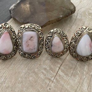 May include: Four silver rings with pink opal stones set in ornate filigree designs. The rings are arranged in a row on a wooden surface.
