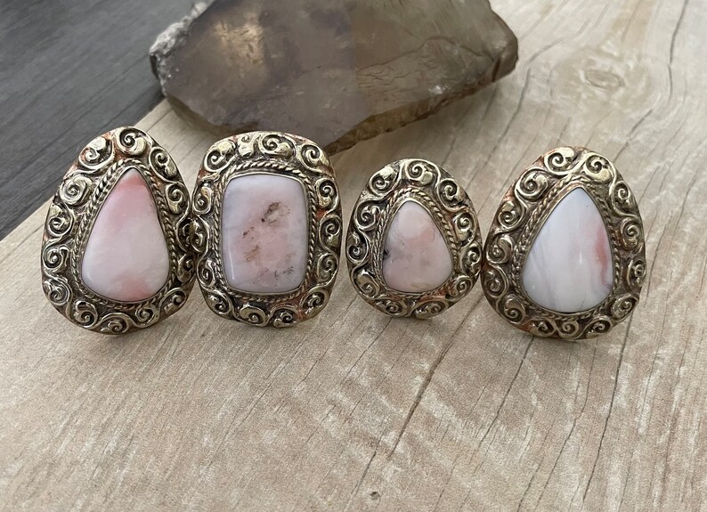 May include: Four silver rings with pink opal stones set in ornate filigree designs. The rings are arranged in a row on a wooden surface.