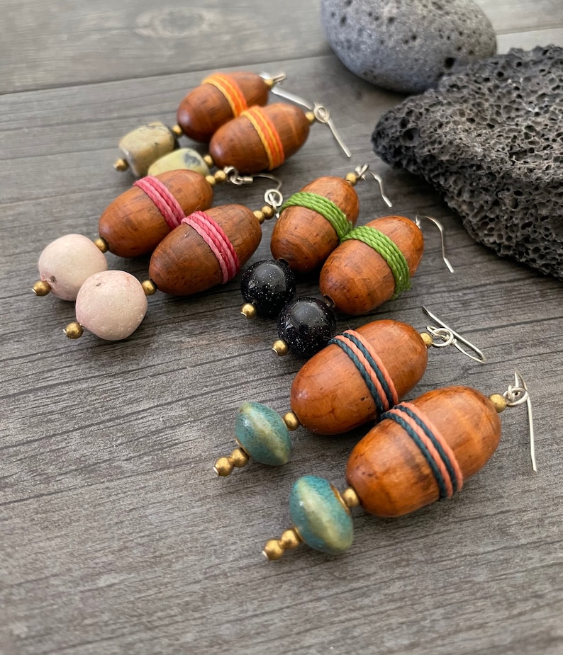 May include: Handmade wooden earrings with a variety of bead accents. The earrings feature brown, oval-shaped wooden beads wrapped with colorful thread, paired with round, speckled, and blue-green beads. The earrings have silver-colored hooks.