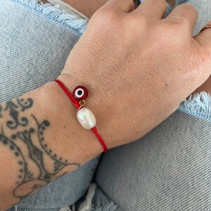 K&ouml;nnte beinhalten: Ein rotes Armband mit einer wei&szlig;en Perle und einem roten b&ouml;sen Blick-Anh&auml;nger.