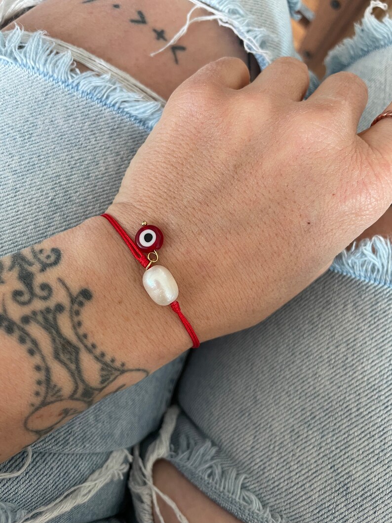 K&ouml;nnte beinhalten: Ein rotes Armband mit einer wei&szlig;en Perle und einem roten b&ouml;sen Blick-Anh&auml;nger.
