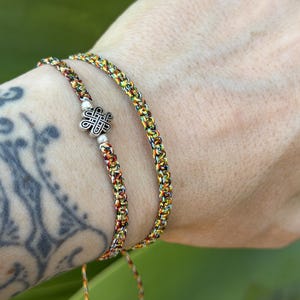 May include: Two braided bracelets made of multicolored thread in shades of yellow, orange, green, and black. One bracelet features a central silver-toned metal charm shaped like an intricate Celtic knot, flanked by two small silver-toned beads.