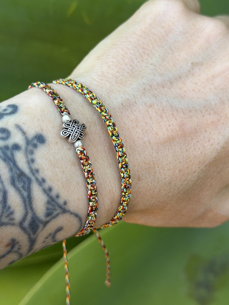May include: Two braided bracelets made of multicolored thread in shades of yellow, orange, green, and black. One bracelet features a central silver-toned metal charm shaped like an intricate Celtic knot, flanked by two small silver-toned beads.
