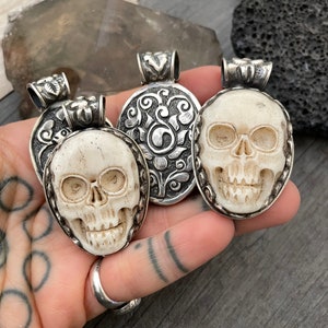 May include: Three silver skull pendants with intricate designs. The skulls are carved from bone and have a white colour. The pendants are on a hand with tattoos.
