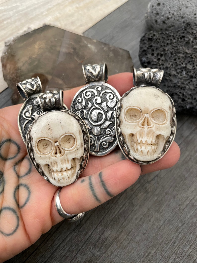 May include: Three silver skull pendants with intricate designs. The skulls are carved from bone and have a white colour. The pendants are on a hand with tattoos.